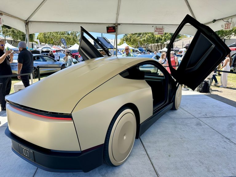 Gold Tesla Cybercab parked outdoors at an event, solid disc wheel covers and butterfly-door silhouette visible, no steering wheel