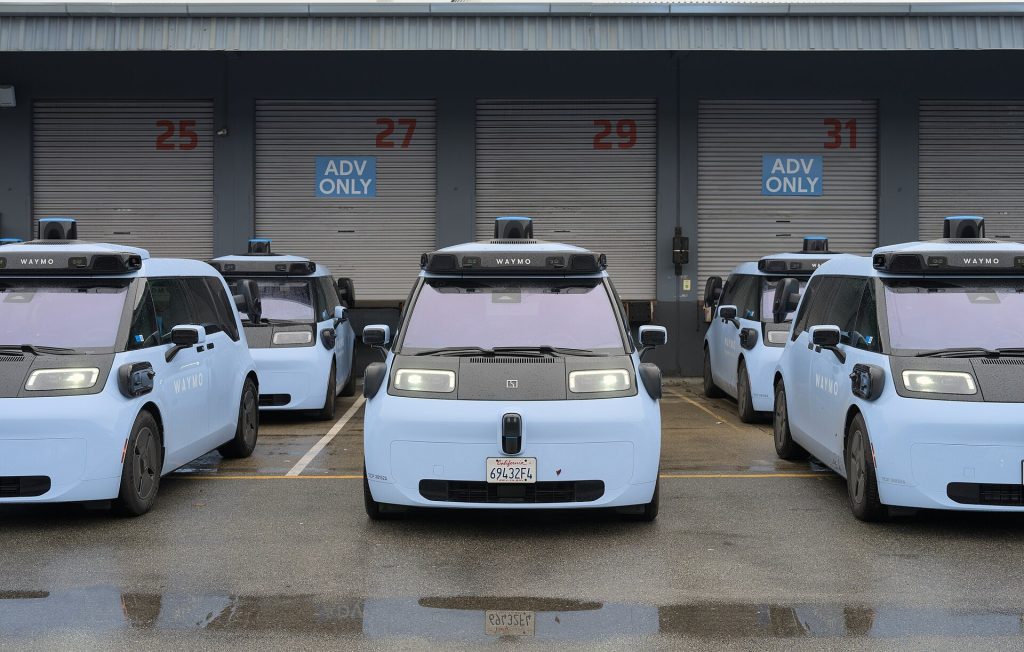 A row of light-blue Waymo Ojai robotaxis with roof sensor domes parked in front of numbered loading bays at a Waymo depot